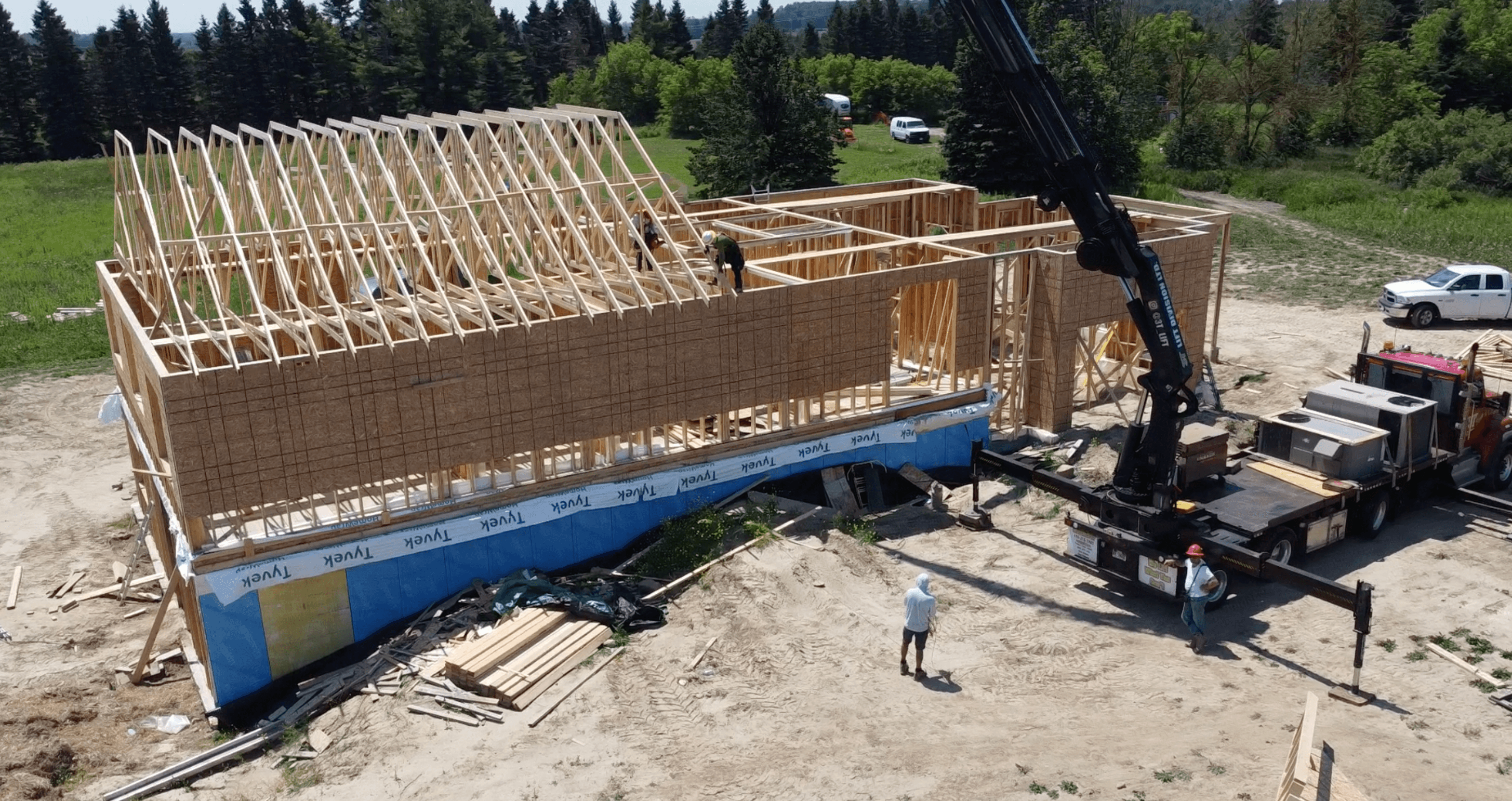 Construction workers install roof trusses on a wooden house frame using a large crane truck.