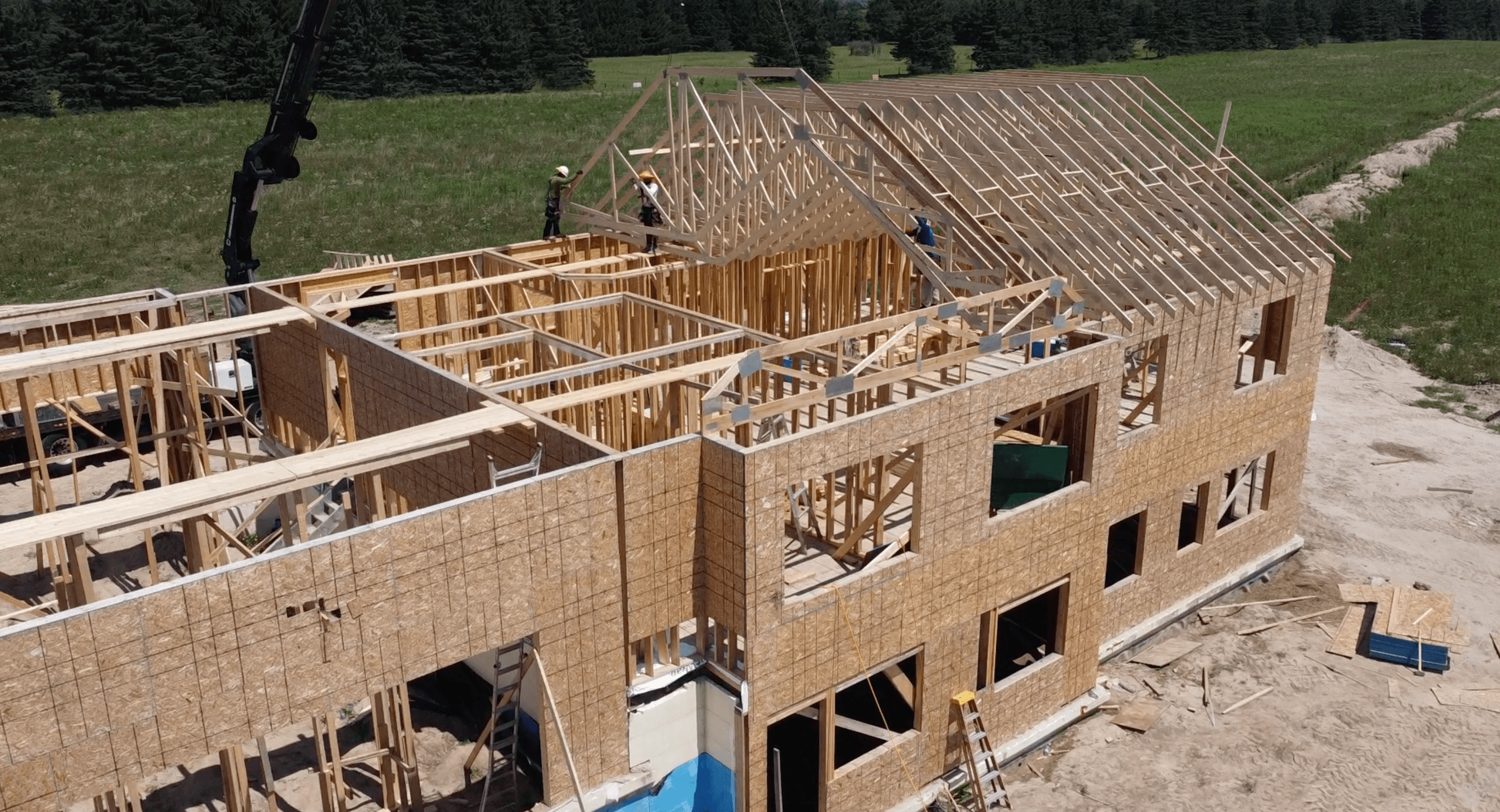 Construction workers install wooden roof trusses on a two-story house frame using a crane.