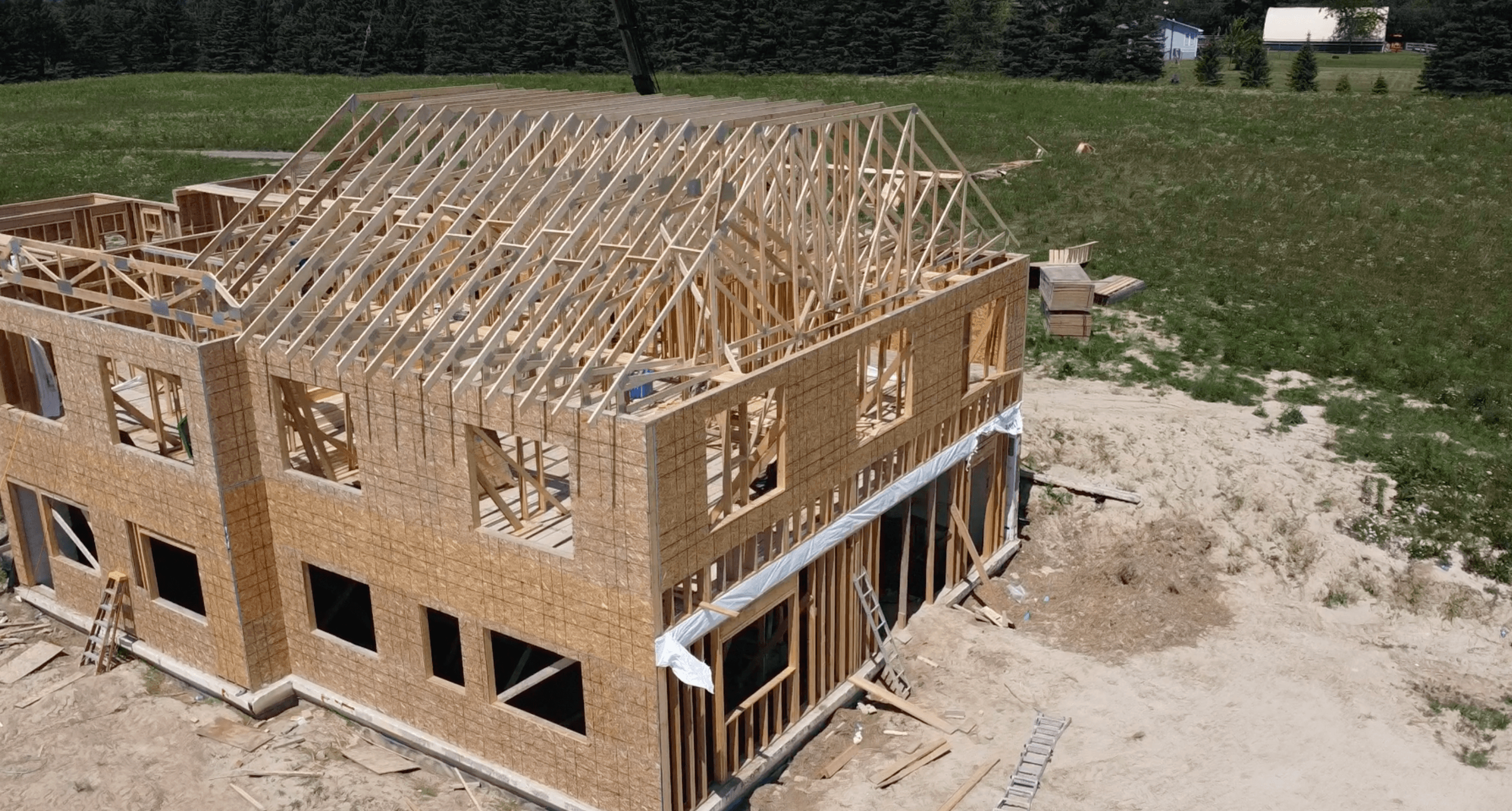 Two-story house under construction with wooden framing and roof trusses in a grassy field.